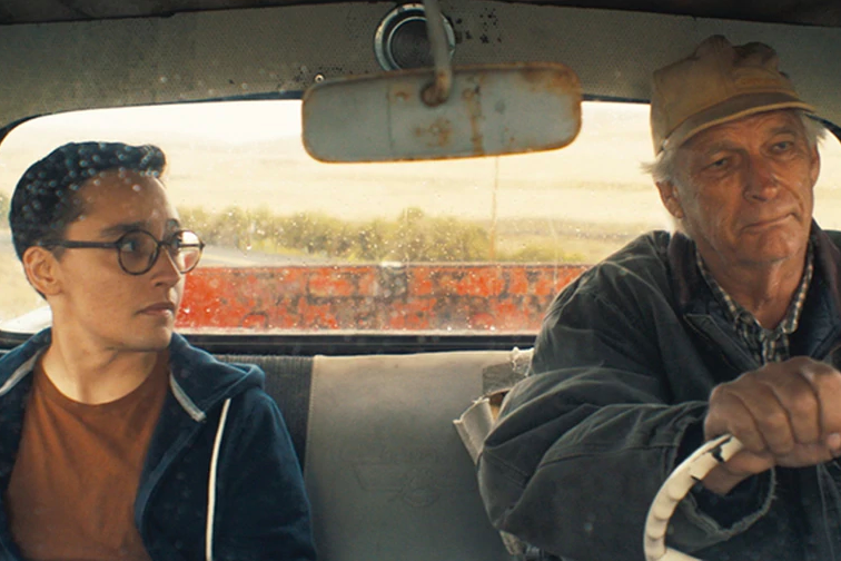 A young man on the left and older man on the right are seated in the passenger and driver seats of an old truck, looking at each other.